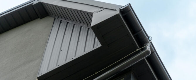 Upward view of a house corner showing black metal roofing, gutters, fascias and soffits against a clear sky.